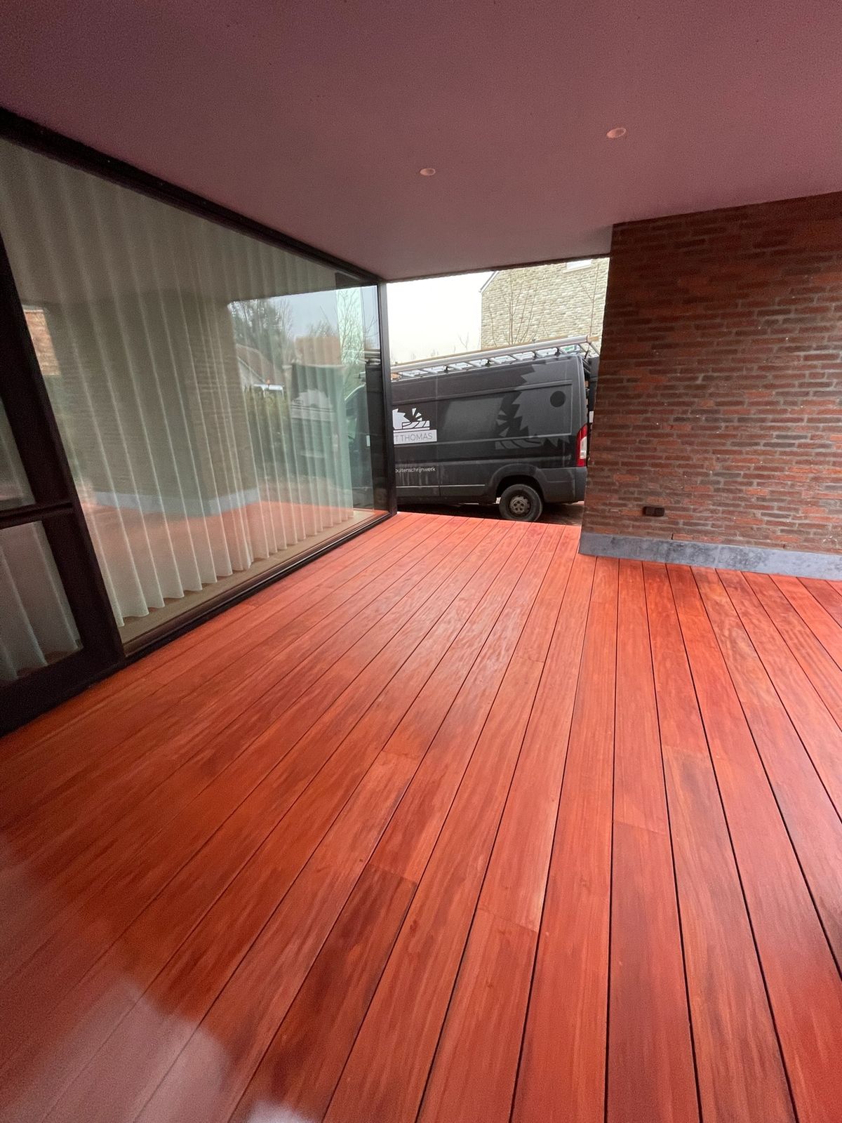 Glanzende hardhouten terrasplanken onder de overkapping langs de schuiframen van een woning in Bazel.