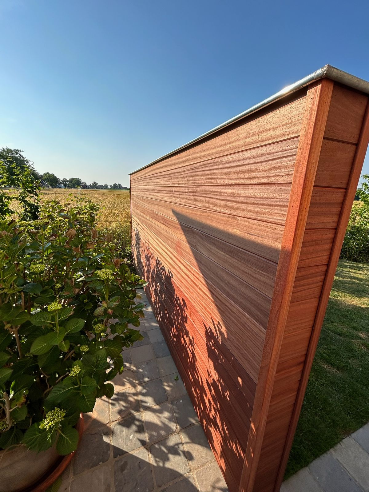 Strak hardhouten tuinscherm met afdekkap tussen tuin en veld in Lotenhulle.
