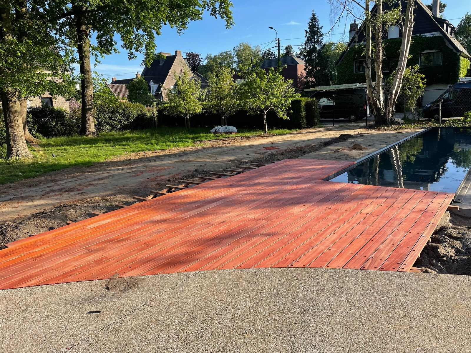 Hardhouten zwembadterras in een grote tuin met bomen en woningen in De Pinte.