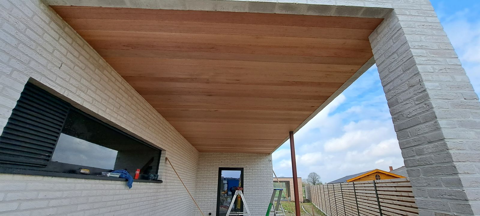 Houten plafond onder een overkapping tegen een witte bakstenen gevel in Torhout.