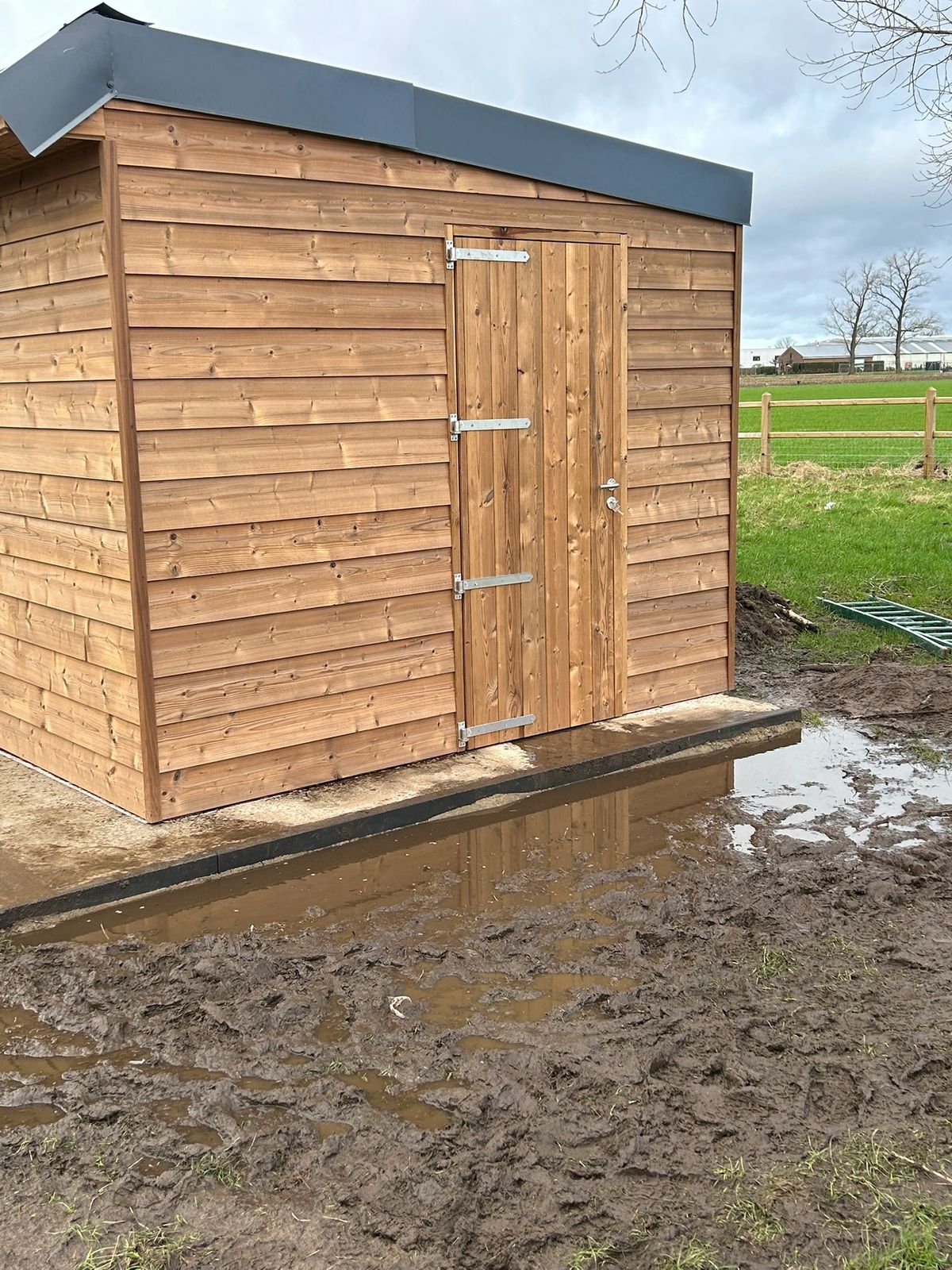 Houten stal met houten deur en plat dak in een modderige weide in Lotenhulle.