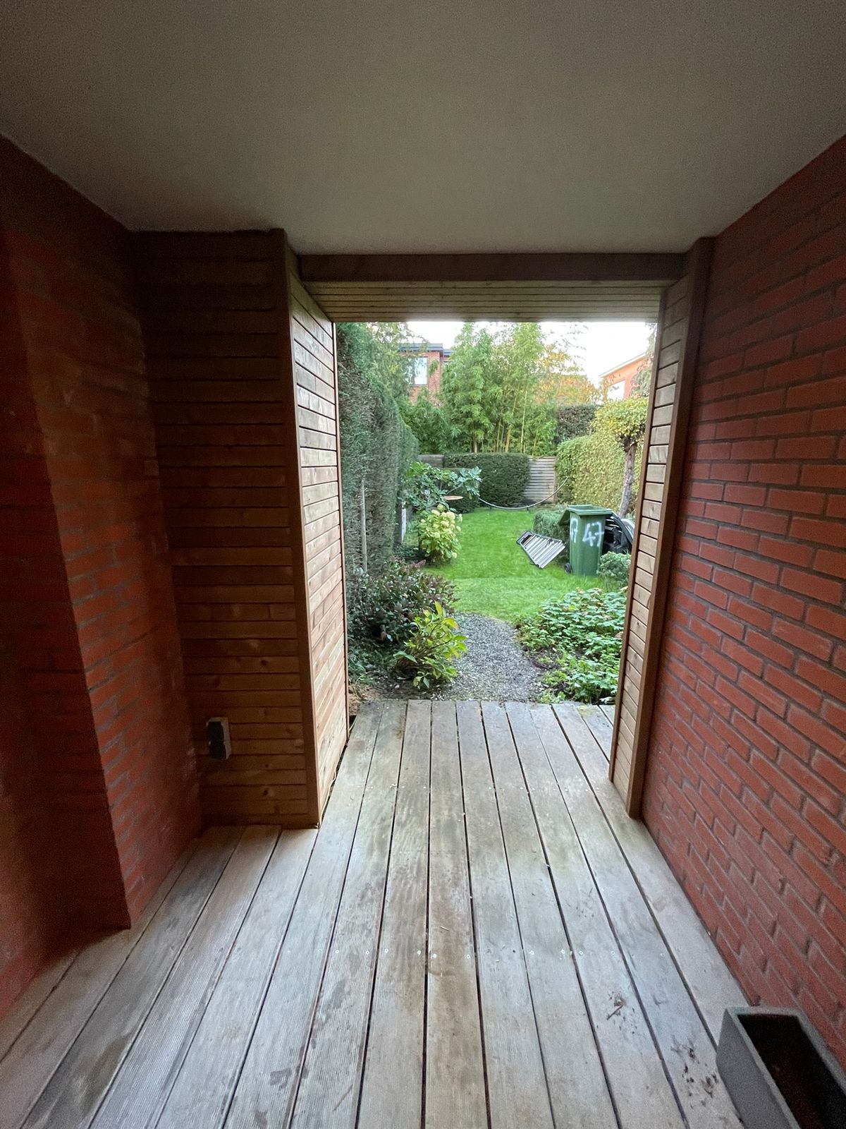 Overdekte doorgang met houten vloer en houten bekleding met zicht op de tuin.