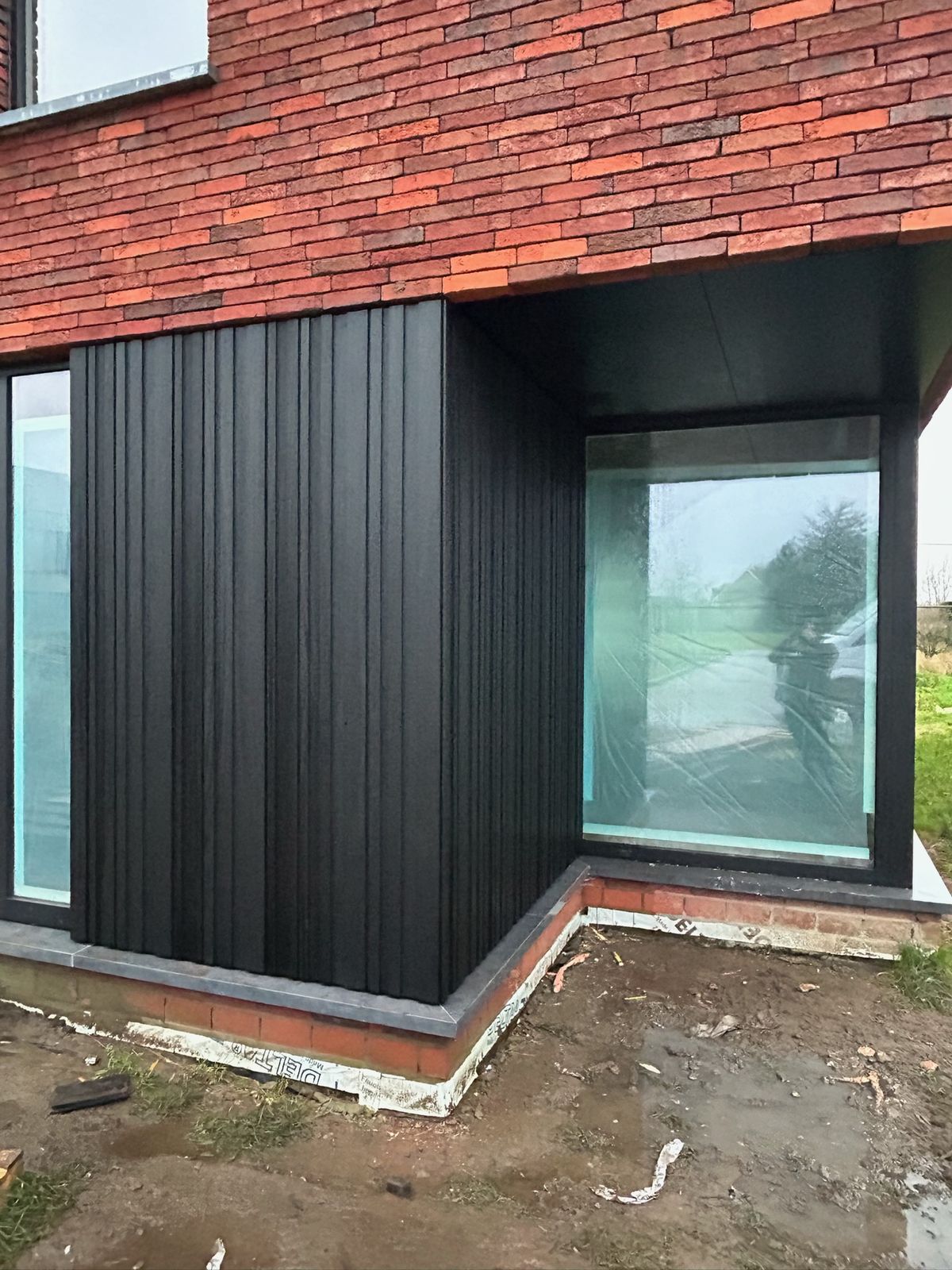 Zwarte gevelbekleding rond een hoekraam onder een overkraging in rode baksteen in Wingene.