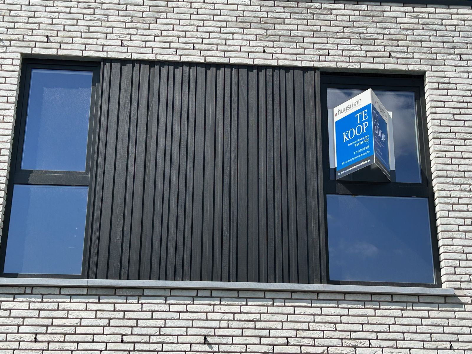 Zwarte houten gevelbekleding tussen twee ramen in een grijze bakstenen gevel in Kortemark.