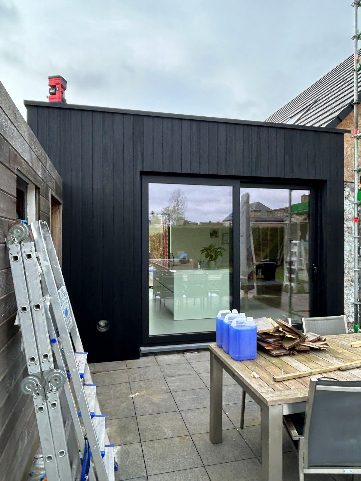 Zwarte houten aanbouw met glazen schuifraam en terras met tuintafel in Teralfene.