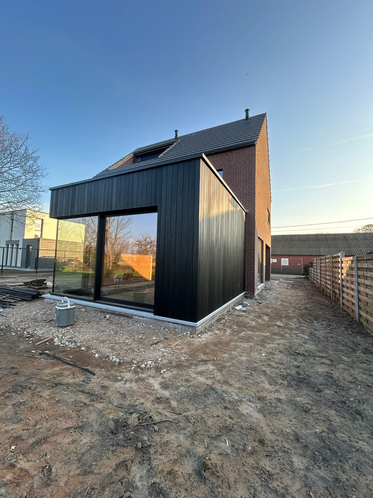 Zijaanzicht van een zwarte houten aanbouw met hoekraam tegen een bakstenen woning in Maldegem.