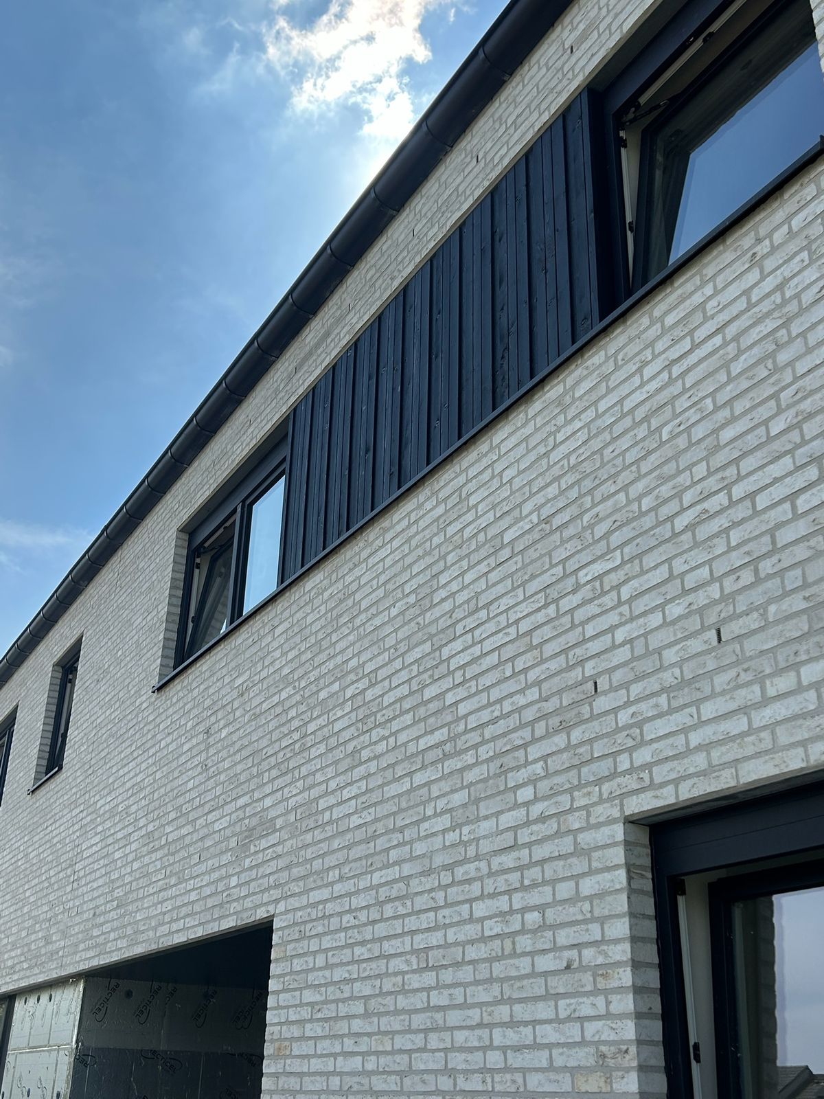 Witte bakstenen woning met zwarte houten gevelbekleding tussen de ramen op de verdieping in Rieme.