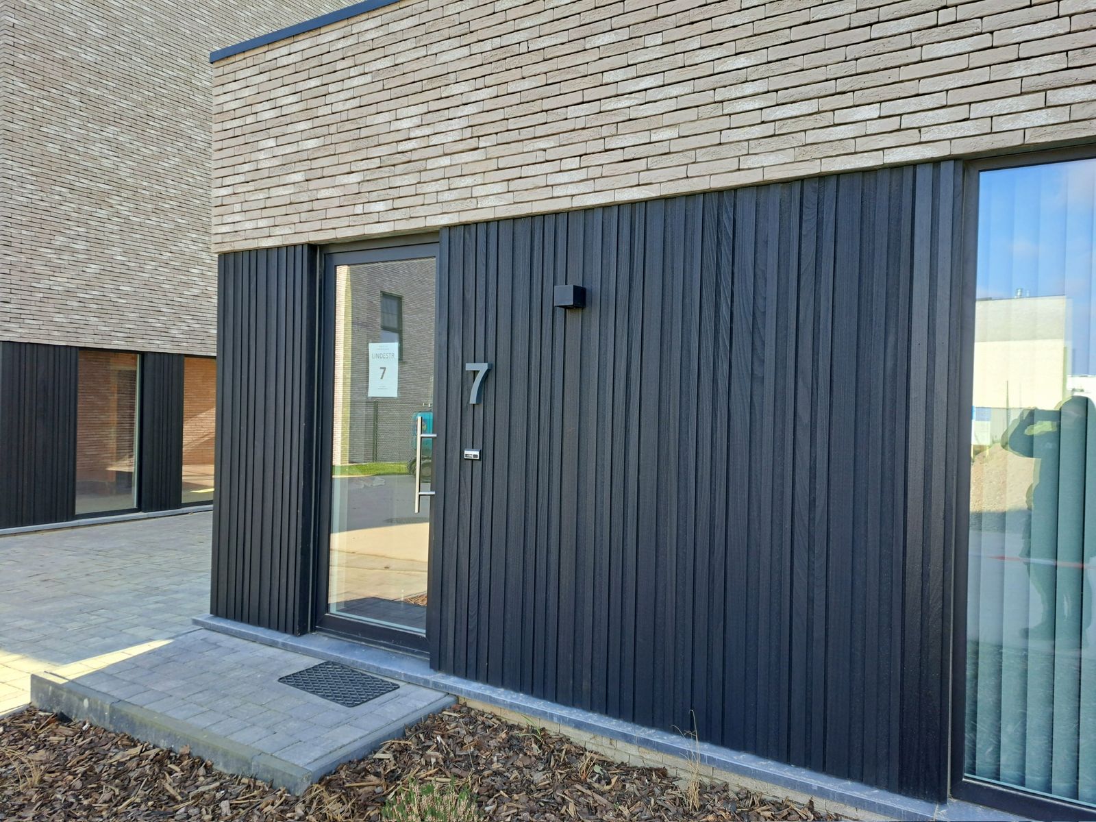 Zwarte houten gevelbekleding naast de voordeur met huisnummer van een woning in Herzele.