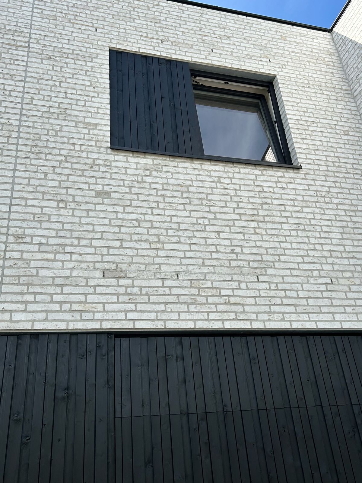 Zwarte houten gevelbekleding onder en naast een raam in een witte bakstenen muur in Rieme.