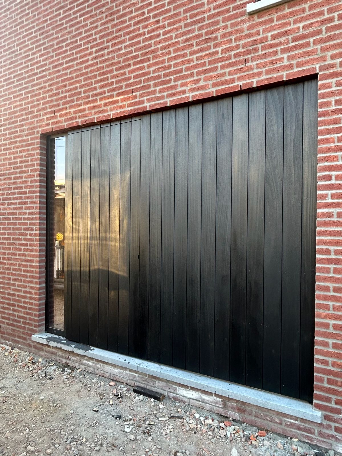 Zwarte houten bekleding in verticale planken naast een grote raamopening in Maldegem.